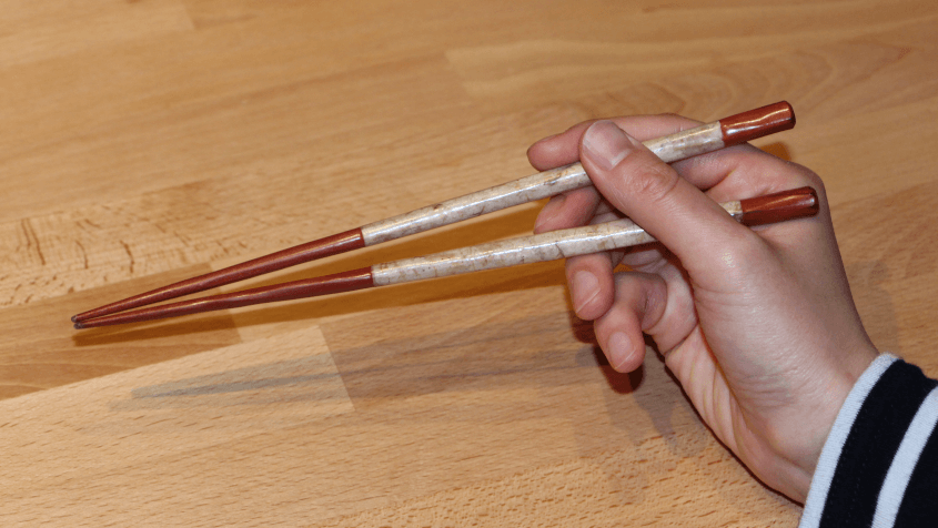 The upper chopstick is placed with its rear at the base of the index finger and with its middle just bellow the fingernail of the middle finger. The top of the thumb fixates it.